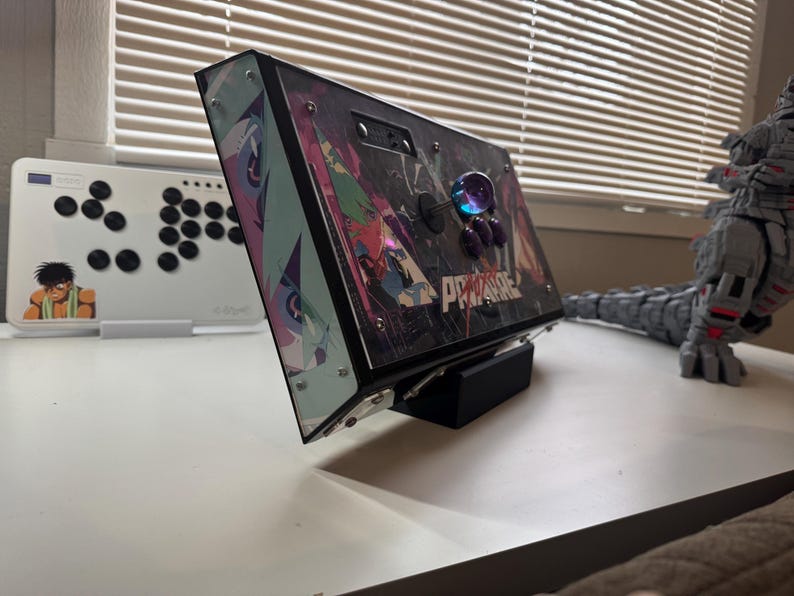 Pu&ograve; includere: Un joystick da arcade nero con un design personalizzato che presenta personaggi anime e la parola "PRIMARE". Il joystick &egrave; su un supporto nero e ha una parte superiore in plastica trasparente.
