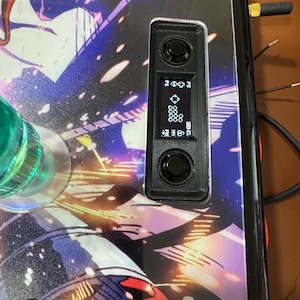 以下が含まれることがあります： カラフルなグラフィックデザインの黒いアーケードコントローラーのクローズアップ。 コントローラーには、アイコンが表示された小さな画面と2つの黒いボタンがあります。 左側には緑色のジョイスティック、右側には金色のアンテナが見えます。