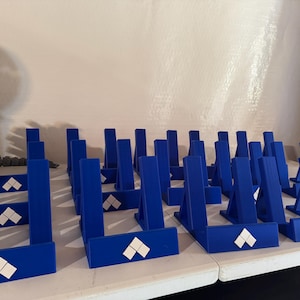 Puede incluir: Un conjunto de expositores azules con un diseño en forma de diamante blanco. Los expositores están dispuestos sobre una superficie blanca, probablemente para exhibir artículos. Están hechos de un material sólido y tienen la misma forma.