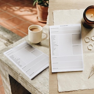 Könnte beinhalten: Zwei weiße Tagesplaner-Blätter mit deutschem Text, eine Tasse Kaffee, eine brennende Kerze in einem braunen Glas und ein paar Perlenringe sind auf einer hellen Oberfläche angeordnet. Die Szene spielt in hellem, natürlichem Licht.