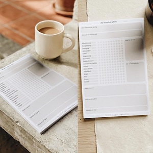 Könnte beinhalten: Zwei weiße Tagesplaner-Blöcke mit grauem Text und Punktrastern werden angezeigt. Eine Tasse Kaffee steht neben einem Block. Eine brennende Kerze in einem braunen Glas, ein paar Perlenringe und Trockenblumen sind ebenfalls zu sehen.