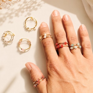Könnte beinhalten: Eine Sammlung von goldfarbenen Drahtringen mit kleinen Perlen in verschiedenen Farben, darunter Weiß, Rosa, Rot und Hellblau. Einige Ringe werden an den Fingern getragen, während andere separat angeordnet sind.