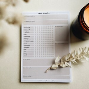 Könnte beinhalten: Ein weißes, druckbares Arbeitsblatt mit dem Titel "Du bist nicht allein." Das Arbeitsblatt enthält eine Liste von Emotionen und ein Raster mit Kreisen zum Verfolgen von Gefühlen. Das Arbeitsblatt liegt auf einer hellbraunen Oberfläche mit einer brennenden Kerze und einer getrockneten Pflanze.