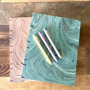 May include: Two notebooks with marbled covers, one pink and one blue-green, with three pens on top. The pens are yellow, pink, and light blue.