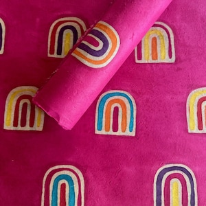 May include: Bright pink paper with a repeating pattern of colorful rainbows. A roll of the same paper is partially visible, showing the texture and design. The rainbows feature red, orange, yellow, blue, and purple arches.
