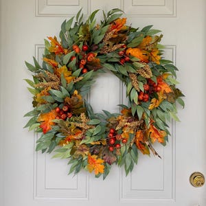 May include: A circular wreath featuring a dense arrangement of green eucalyptus-style leaves, vibrant orange and yellow autumn maple leaves, clusters of small red berries, and textured golden-brown wheat stalks, all mounted on a light-colored door.