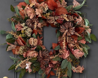 May include: A circular autumn wreath featuring a dense arrangement of dried oak leaves in shades of rust, copper, and brown, interspersed with small brown decorative pumpkins, dried flower clusters, and green foliage on a dark gray door.