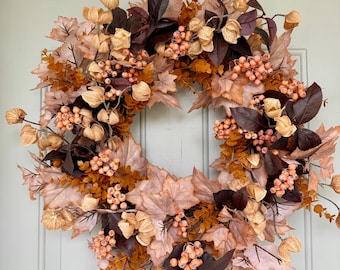 May include: A circular wreath featuring a mix of autumn-toned foliage, including light brown maple leaves, dark brown leaves, clusters of small peach-colored berries, and dried papery husks in a soft beige hue, arranged in a dense, layered circular pattern.