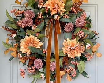 May include: A circular wreath featuring large peach-colored dahlia flowers, smaller dusty pink pom-pom blooms, green leaves, and clusters of small orange berries. A long, vertical, dark orange velvet ribbon hangs from the center of the wreath.