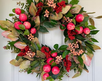 May include: A circular wreath featuring artificial red apples, clusters of bright red berries, and small pale orange berries. The arrangement is accented with dark green leaves and variegated foliage in shades of green, yellow, and soft pinkish-brown.