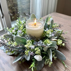 May include: A lit candle in a glass jar, surrounded by a wreath of green and gray foliage, small purple flowers, and seashells. The candle sits on a wooden table near a window.