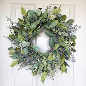 May include: A circular wreath composed of various green leaves, including eucalyptus and other foliage, with accents of lavender and small white flowers. The wreath is displayed against a white door.