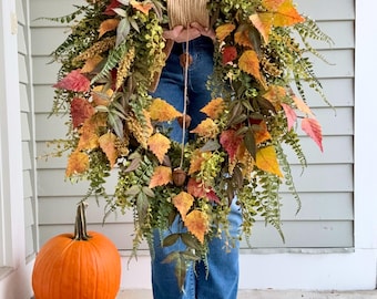 May include: A teardrop-shaped autumn wreath featuring a mix of green ferns, yellow and orange maple leaves, and small brown acorns. The wreath is constructed with a central jute twine hanger and displays a vibrant array of fall foliage colors.