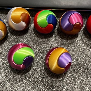 Könnte beinhalten: Sieben bunte, kugelförmige Fidget-Spielzeuge mit einem wirbelnden Muster. Jedes Spielzeug besteht aus drei verschiedenen Farben, wodurch ein einzigartiges Design entsteht.
