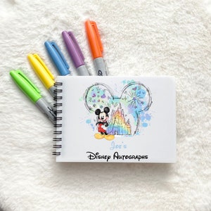 May include: A white spiral bound notebook with a colourful watercolour illustration of Mickey Mouse and Cinderella's castle. The notebook has the text "Joe's Disney Autographs" printed on the front.