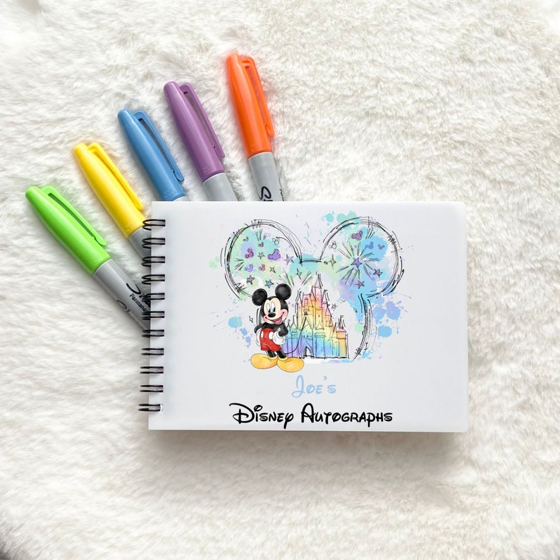 May include: A white spiral bound notebook with a colorful watercolor illustration of Mickey Mouse and Cinderella's castle. The notebook has the text "Joe's Disney Autographs" printed on the front.