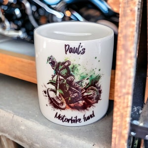 May include: White ceramic money bank with a slot at the top. The bank features a graphic of a motocross rider in green and black, with the text "Paul's Motorbike fund".