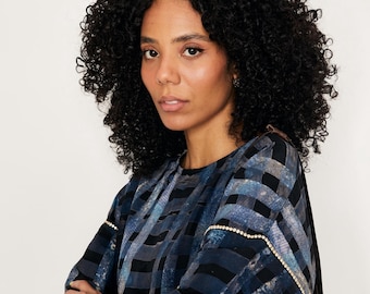 Può includere: Una donna con i capelli neri ricci indossa una camicia a fantasia blu e nera con un bordo di strass. La camicia ha una vestibilità rilassata e una silhouette leggermente oversize.