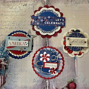 Pode incluir: Quatro enfeites de artesanato de papel em vermelho, branco e azul com temas patrióticos. Os enfeites apresentam as palavras "America", "Liberty", "Freedom" e "Let's Celebrate".