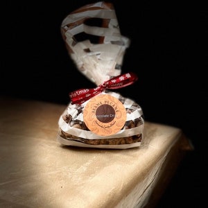 May include: A clear plastic bag filled with chocolate-covered pecans. The bag is tied with a red ribbon with white snowflakes and the words "Merry Christmas". A white circular label with black text reads "Sean's Pecans Chocolate Dreams Handmade Candies & Treats".