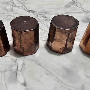 May include: Four copper hexagonal objects with varying patinas. The objects are arranged in a row, with the copper tones ranging from dark brown to bright copper. The objects are on a marble-patterned surface.