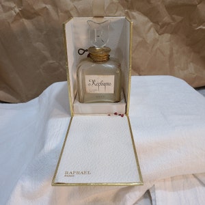 May include: Vintage perfume bottle in a cream-colored box. The bottle is square with a floral-shaped stopper and the word "Replique" in script. The box is open, revealing the bottle inside. The box has gold trim and the words "Raphael Paris" are printed on the bottom.
