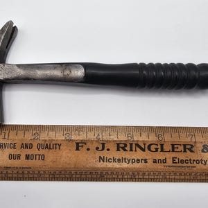 Puede incluir: Un martillo de uña con mango negro y cabeza de metal, junto a una regla de madera. La regla está marcada en centímetros y tiene el texto "SERVICE AND QUALITY OUR MOTTO" y "F. J. RINGLER & CO."