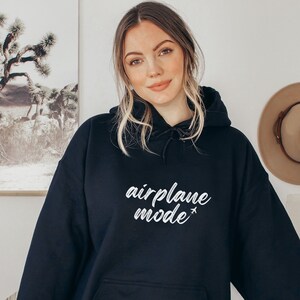 May include: A black hooded sweatshirt with the words "airplane mode" in white script. The sweatshirt has a front pocket and a drawstring hood. The model is wearing the sweatshirt.