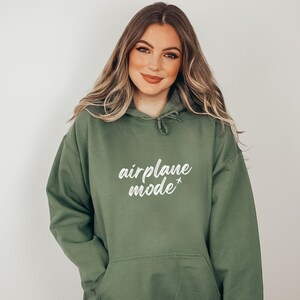 May include: Olive green hoodie with the text "airplane mode" in white script. The hoodie has a drawstring hood and a front pocket. The model is wearing the hoodie.