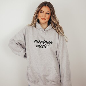 May include: A light gray hoodie with the words "airplane mode" in black script. The hoodie has a drawstring hood and a front pocket. The text includes a small airplane symbol. The person in the image is wearing the hoodie.
