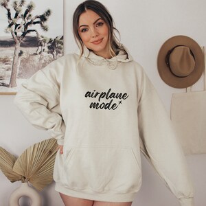 May include: Cream-colored hoodie with the text "airplane mode" in black script. The hoodie has a drawstring hood and a front pocket. A casual, comfortable garment for everyday wear.