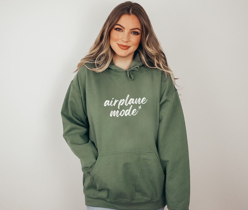 May include: Olive green hoodie with the text "airplane mode" in white script. The hoodie has a drawstring hood and a front pocket. The model is wearing the hoodie.