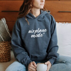 May include: A dark gray hoodie with the words "airplane mode" in white script, accompanied by a small airplane graphic. The hoodie has a front pocket and a hood. The person is holding a white mug.