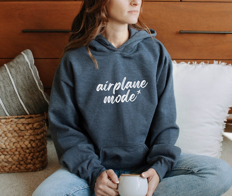 May include: A dark gray hoodie with the words "airplane mode" in white script, accompanied by a small airplane graphic. The hoodie has a front pocket and a hood. The person is holding a white mug.