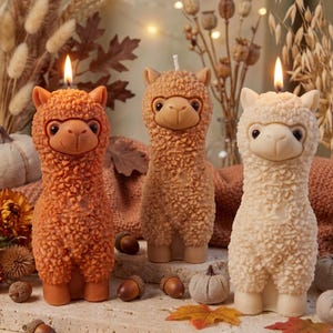 May include: Three llama-shaped candles in orange, tan, and cream colours. Each candle features a textured, wool-like surface, a smooth face with dark eyes, and a small nose. Two of the candles are lit with a visible flame at the top of the head.