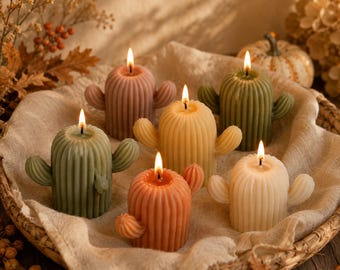 May include: A collection of five cactus-shaped candles in muted shades of pink, sage green, yellow, orange, and cream. Each candle features a ribbed texture, two side arms, and a lit wick, arranged together in a woven basket lined with fabric.