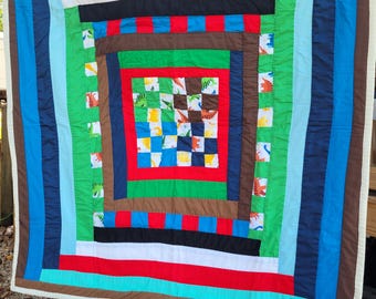 May include: A colorful handmade quilt with a square-in-square design. The quilt features a variety of colors including red, blue, green, brown, and black. The center square has a checkerboard pattern, and the surrounding squares have dinosaur patterns.