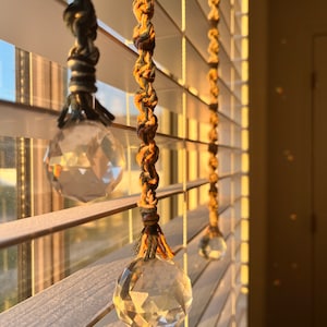 May include: Three crystal prisms hang from macrame cords in front of a white wooden window blind. The prisms are catching the light and creating rainbows.