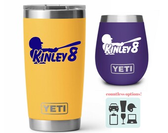 May include: Two insulated tumblers, one yellow and one purple, each with a baseball-themed design. The yellow tumbler has a blue graphic with the name "KINLEY 8". The purple tumbler has the same design. Both have the "YETI" logo.