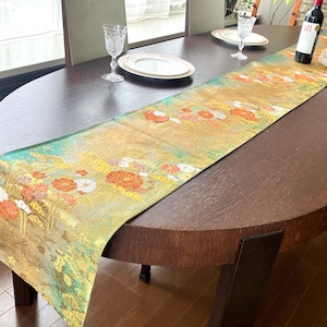 May include: A decorative table runner with a floral pattern in shades of gold, orange, and green. The runner is placed on a dark brown oval dining table. Wine glasses, plates, and a wine bottle are also on the table.