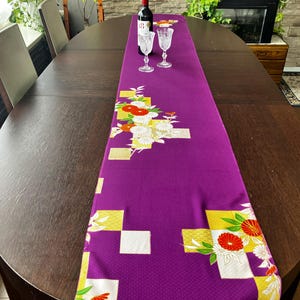 Puede incluir: Un camino de mesa rectangular de color morado con un patrón repetitivo de cuadrados geométricos blancos y dorados, acentuado con motivos florales en rojo, blanco y naranja. La tela presenta un tejido fino y sutil en todo el diseño.