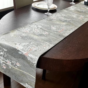 May include: A long, rectangular table runner featuring a light grey and silver base with an intricate woven pattern of floral motifs and geometric shapes. The fabric displays subtle accents of soft pink and white throughout the detailed design.