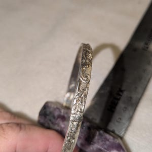 May include: A silver-toned metal bangle bracelet featuring an intricate embossed floral pattern along its outer surface. The bracelet is positioned next to a metal ruler showing a measurement of approximately 2.5 inches in diameter.