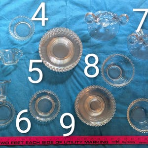 Puede incluir: Una colección de fuentes y piezas decorativas de vidrio transparente, que incluyen cuencos pequeños, un azucarero y una bandeja de servir. Las piezas están dispuestas sobre una superficie azul, con una regla para la escala. Las piezas tienen bordes decorativos.