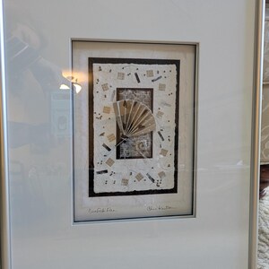 May include: Framed artwork featuring a fan design. The artwork has a white background with a rectangular border and a silver frame. The fan is made of beige and brown paper. The artwork is signed and titled "Confetti Fan."