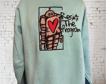 May include: A light blue sweatshirt featuring a robot design with a red heart on its chest, framed in a black rectangle. The words "Resist The Program" are written in black script to the right of the robot.