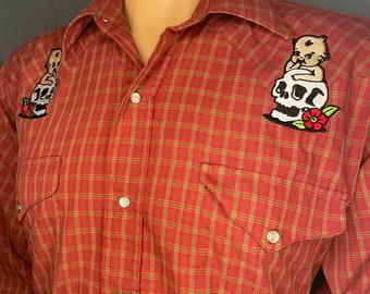 May include: Red and green plaid long-sleeved button-up shirt with two embroidered patches of a cherub holding a skull on each shoulder.