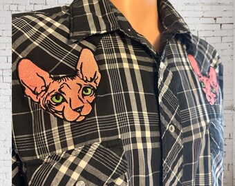 May include: Black and white plaid button-up shirt with two pink Sphynx cat patches. The cat patches have green eyes and are on the chest and upper right shoulder. The shirt has two chest pockets and silver buttons.