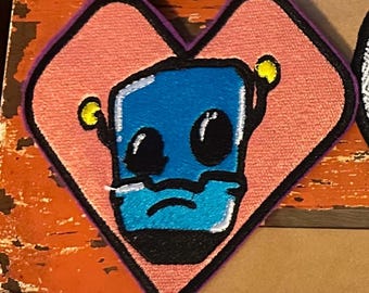 May include: Embroidered patch in the shape of a heart with a purple border. The patch features a blue robot face with large black eyes, a black mouth, and yellow antenna-like details. The background is peach-colored.