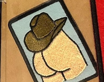 May include: Embroidered patch featuring a cowboy hat over a stylized depiction of a rear end. The patch has a light blue background with a black border. The hat is brown and the rear end is tan.